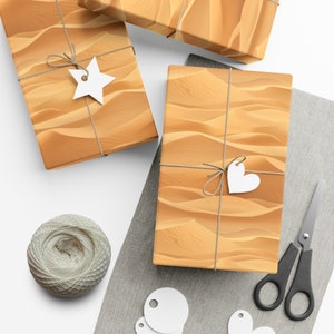 Pode incluir: Três caixas de presente embrulhadas em papel castanho com um padrão de dunas de areia do deserto. As caixas são amarradas com barbante e têm etiquetas de papel branco com a forma de uma estrela, coração e círculo.