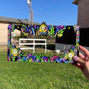 May include: A black license plate frame with a colorful psychedelic design featuring mushrooms, a monster, and dripping paint. The design is in shades of green, yellow, pink, purple, and blue.