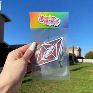 May include: A clear plastic bag with a black string hanging from the top. Inside the bag is a white diamond-shaped sticker with red and black marbled design and the word "SLANDER" in white letters. The bag has a colorful label with the text "Karma Kandi" on it.
