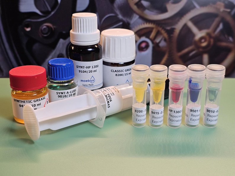 May include: A collection of watchmaking lubricants and tools, including small bottles of grease and oil, a syringe, and several vials with colored liquids. The bottles have white caps and labels with text. The background features watch gears.