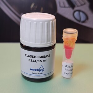 May include: A bottle of Classic Grease 8213/15 ml with a white cap and a small vial containing a pink liquid. The bottle has a black base and white label with the brand name "moebius" and "Swiss Made".