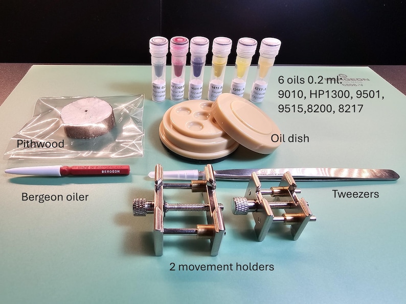 May include: A collection of watch repair tools, including a Bergeon oiler, tweezers, two movement holders, an oil dish, and six vials of oil. Also included is a piece of pithwood. The tools are arranged on a green surface.