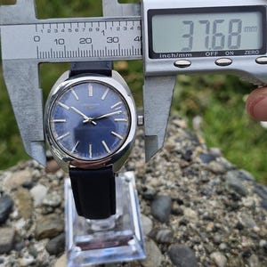 May include: A vintage Longines watch with a navy blue dial and silver hands. The watch has a black leather strap and is being measured with a digital caliper. The watch face measures 37.68 mm.