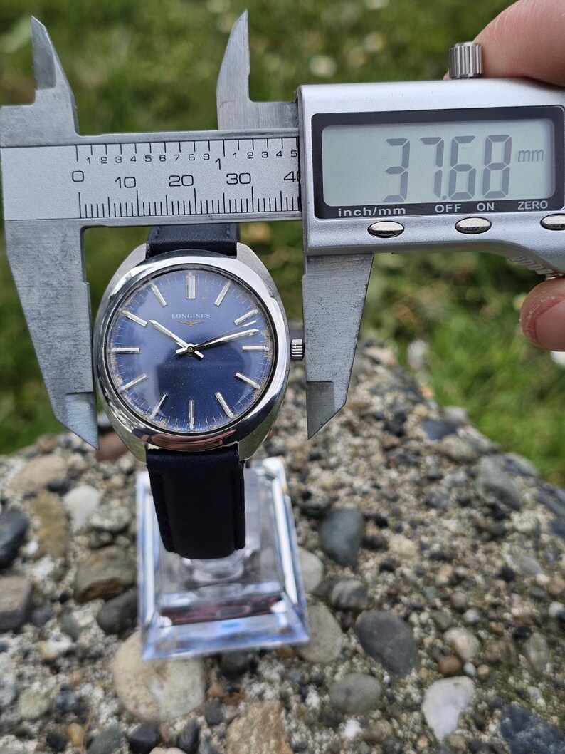 May include: A vintage Longines watch with a navy blue dial and silver hands. The watch has a black leather strap and is being measured with a digital caliper. The watch face measures 37.68 mm.