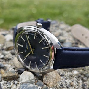 May include: A silver-cased wristwatch with a dark blue face and gold hands, resting on a bed of rocks. The watch has a dark blue leather band. The watch face displays the brand name "Longines".