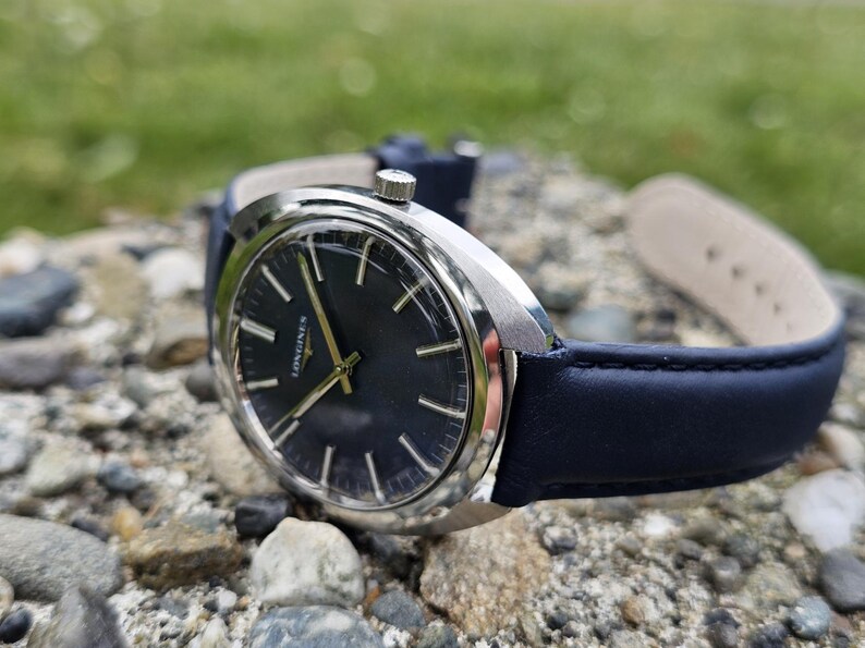 May include: A silver-cased wristwatch with a dark blue face and gold hands, resting on a bed of rocks. The watch has a dark blue leather band. The watch face displays the brand name "Longines".