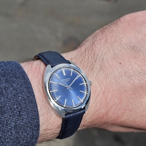May include: A vintage wristwatch with a blue dial and silver-tone case. The watch features a dark blue leather band. The watch face has silver-tone hour markers and hands. The brand name "LONGINES" is printed on the watch face.