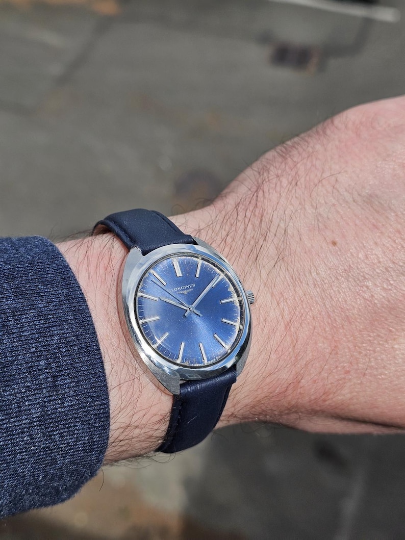 May include: A vintage wristwatch with a blue dial and silver-tone case. The watch features a dark blue leather band. The watch face has silver-tone hour markers and hands. The brand name "LONGINES" is printed on the watch face.