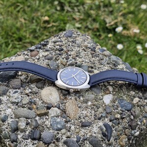 May include: A wristwatch with a blue face and silver-colored case, set against a backdrop of a stone surface and green grass. The watch has a blue leather band and silver buckle. The watch hands are silver.