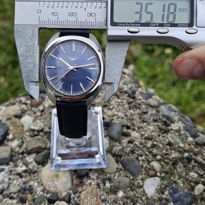 May include: A vintage Longines wristwatch with a dark blue dial and silver hands. The watch is measured with a digital caliper, showing a diameter of 35.18 mm (1.39 inches). It has a black leather strap and is displayed on a clear stand.