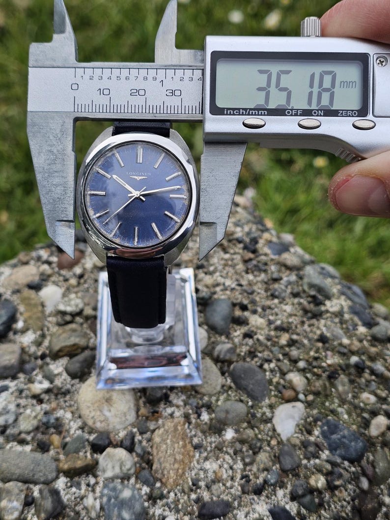 May include: A vintage Longines wristwatch with a dark blue dial and silver hands. The watch is measured with a digital caliper, showing a diameter of 35.18 mm (1.39 inches). It has a black leather strap and is displayed on a clear stand.