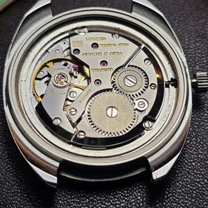 May include: Close-up of a vintage Longines watch movement. The watch mechanism features intricate gears, jewels, and the text "Seventeen 17 Jewels." The watch case is silver-toned, and the movement is set against a dark background.