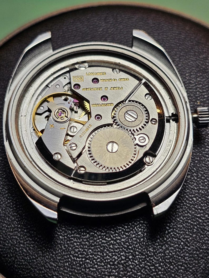 May include: Close-up of a vintage Longines watch movement. The watch mechanism features intricate gears, jewels, and the text "Seventeen 17 Jewels." The watch case is silver-toned, and the movement is set against a dark background.