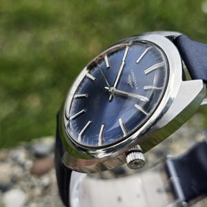 May include: A vintage wristwatch with a dark blue face and silver-colored casing. The watch has silver hands and markers, and a dark blue leather band. The watch face has the word "LONGINES" printed on it.