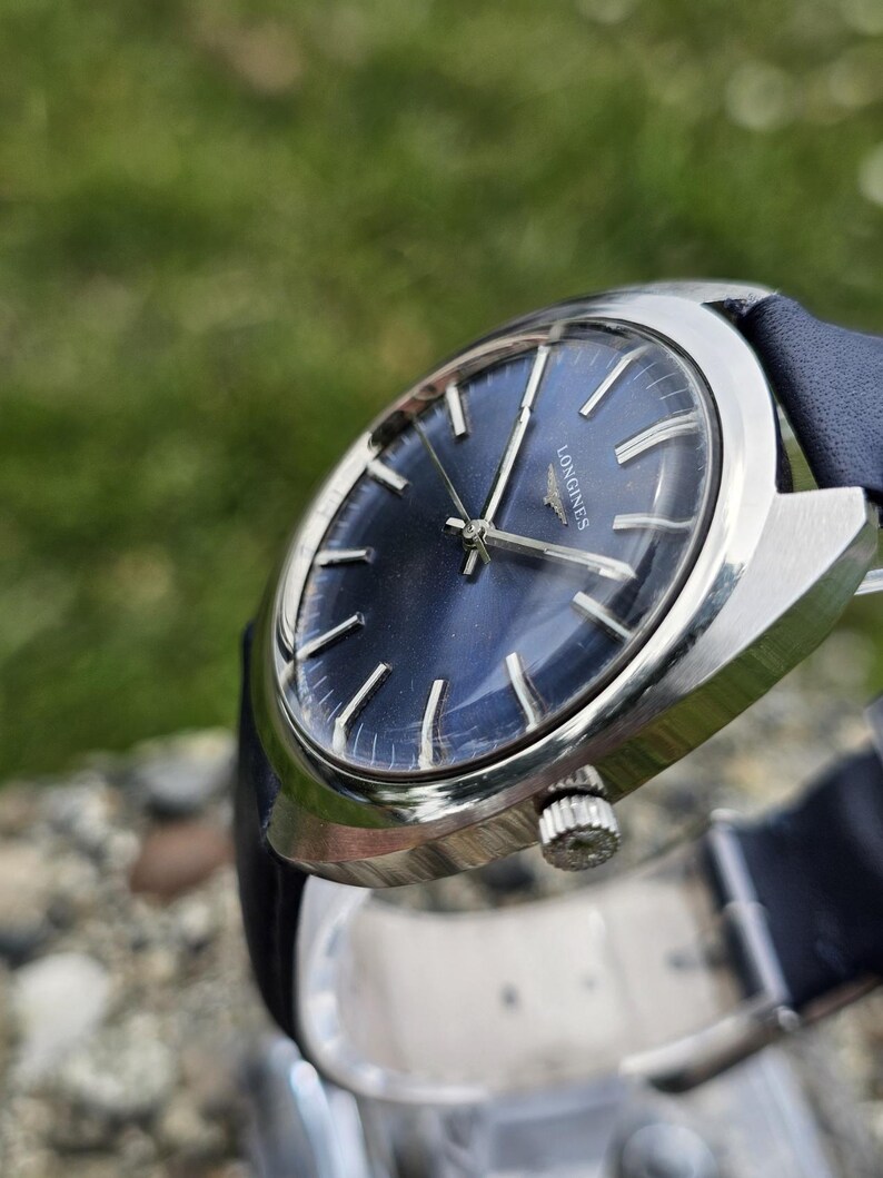 May include: A vintage wristwatch with a dark blue face and silver-colored casing. The watch has silver hands and markers, and a dark blue leather band. The watch face has the word "LONGINES" printed on it.