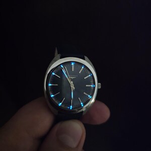 May include: A vintage wristwatch with a dark blue face and silver casing. The watch hands and hour markers glow with a blue luminescence. The watch has a dark blue leather band.
