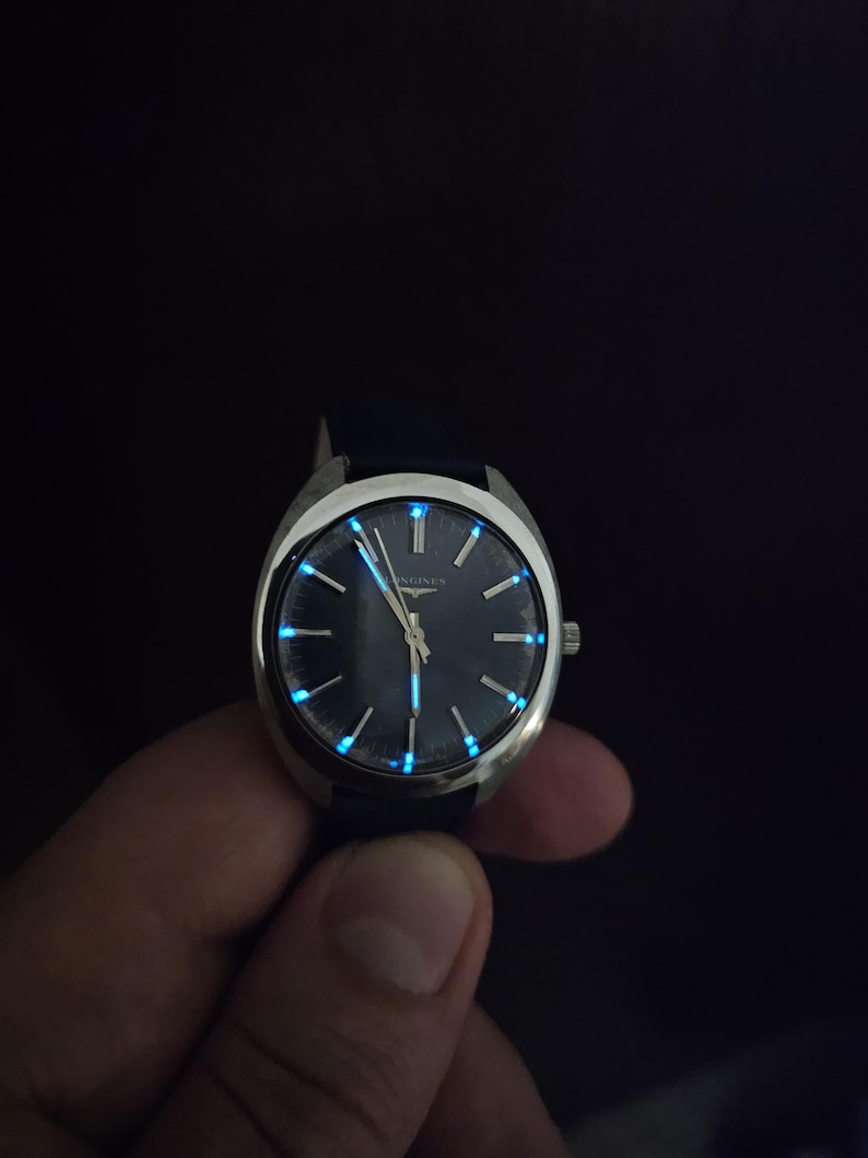 May include: A vintage wristwatch with a dark blue face and silver casing. The watch hands and hour markers glow with a blue luminescence. The watch has a dark blue leather band.