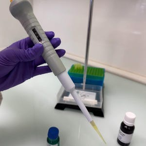 May include: A gray and white pipette with a yellow tip is held by a gloved hand. Two small bottles, one green with a blue cap and one black with a white cap, are on the table. A rack of pipette tips is in the background.
