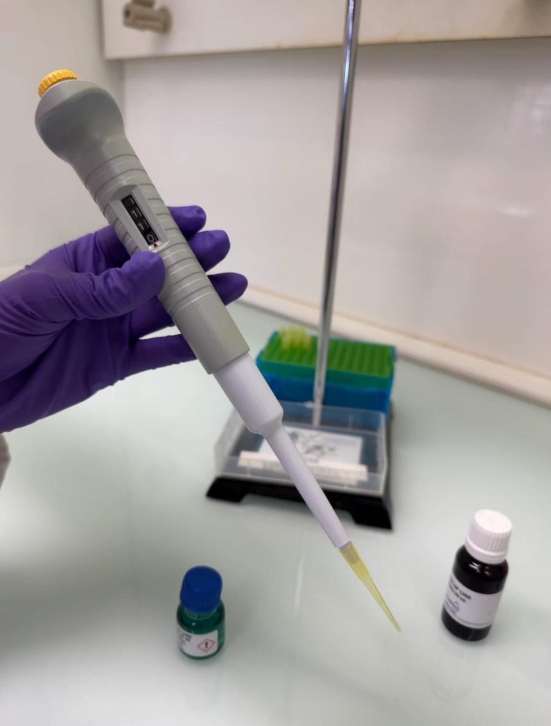 May include: A gray and white pipette with a yellow tip is held by a gloved hand. Two small bottles, one green with a blue cap and one black with a white cap, are on the table. A rack of pipette tips is in the background.