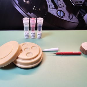May include: A collection of watch repair tools and components, including a beige watch holder, three vials with colored liquids, a red screwdriver, a white tool, and a wooden disc. The background features a watch mechanism.