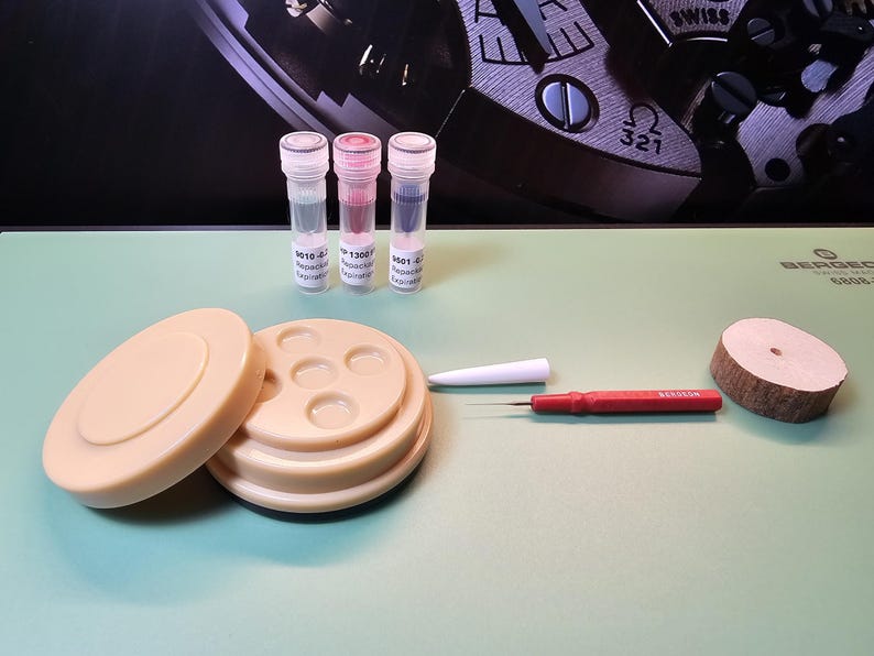 May include: A collection of watch repair tools and components, including a beige watch holder, three vials with colored liquids, a red screwdriver, a white tool, and a wooden disc. The background features a watch mechanism.