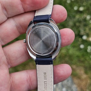 May include: A silver-toned wristwatch with a navy blue leather band. The watch face is visible, with the words "WATER RESISTANT", "ANTIMAGNETIC", and "SHOCK" printed on the back. The band is marked "ECHT LEDER 17".