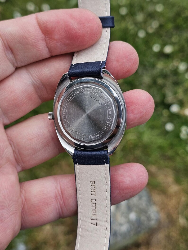 May include: A silver-toned wristwatch with a navy blue leather band. The watch face is visible, with the words "WATER RESISTANT", "ANTIMAGNETIC", and "SHOCK" printed on the back. The band is marked "ECHT LEDER 17".