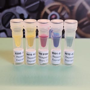 May include: Five small, clear vials with white labels and clear caps, each containing a different colored liquid: yellow, yellow-orange, red-purple, blue, and teal. The vials are arranged in a row against a blurred background.