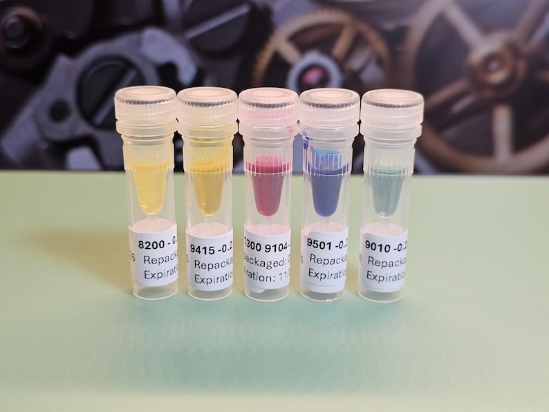 May include: Five small, clear vials with white labels and clear caps, each containing a different colored liquid: yellow, yellow-orange, red-purple, blue, and teal. The vials are arranged in a row against a blurred background.