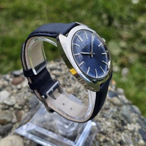 May include: A vintage wristwatch with a dark blue face and silver-colored casing. The watch has a black leather band and is displayed on a clear stand. The watch face has silver-colored hands and markers. The brand name "LONGINES" is visible on the watch face.