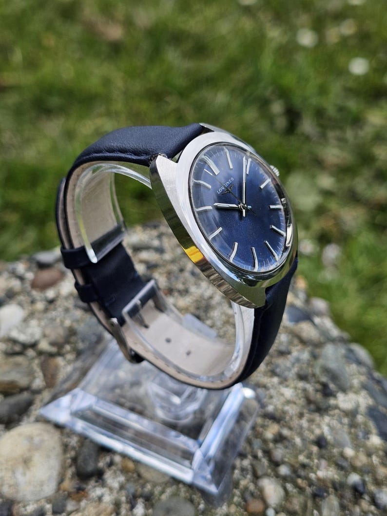 May include: A vintage wristwatch with a dark blue face and silver-colored casing. The watch has a black leather band and is displayed on a clear stand. The watch face has silver-colored hands and markers. The brand name "LONGINES" is visible on the watch face.