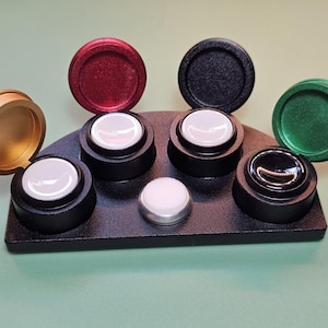 May include: A black semi-circular base holds four circular containers with hinged lids in gold, red, black, and green. Each container features a white interior insert, except for the green one which holds a black insert. A small white button sits in the center.