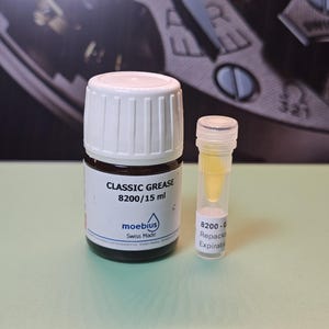 May include: A bottle of "CLASSIC GREASE 8200/15 ml" by Moebius, Swiss Made, next to a small vial of yellow liquid. The bottle has a white cap and a brown label. The background is a watch face.