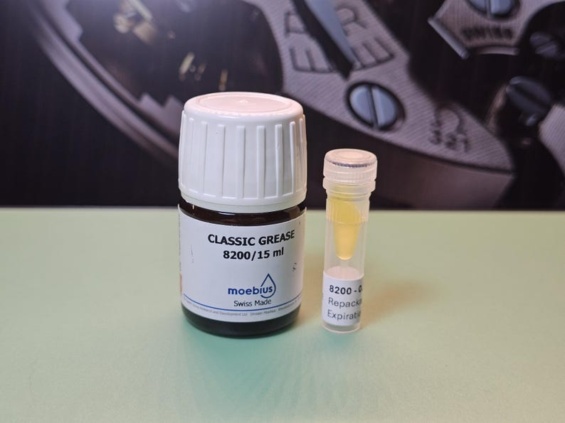 May include: A bottle of "CLASSIC GREASE 8200/15 ml" by Moebius, Swiss Made, next to a small vial of yellow liquid. The bottle has a white cap and a brown label. The background is a watch face.