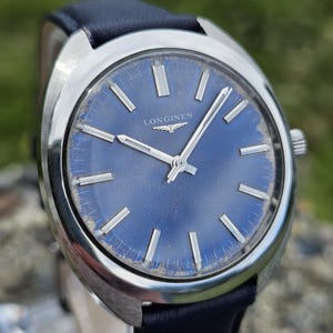 May include: A vintage Longines wristwatch with a blue dial and silver hands and markers. The watch has a stainless steel case and a dark blue leather strap. The dial features the Longines logo and the words "SWISS MADE".
