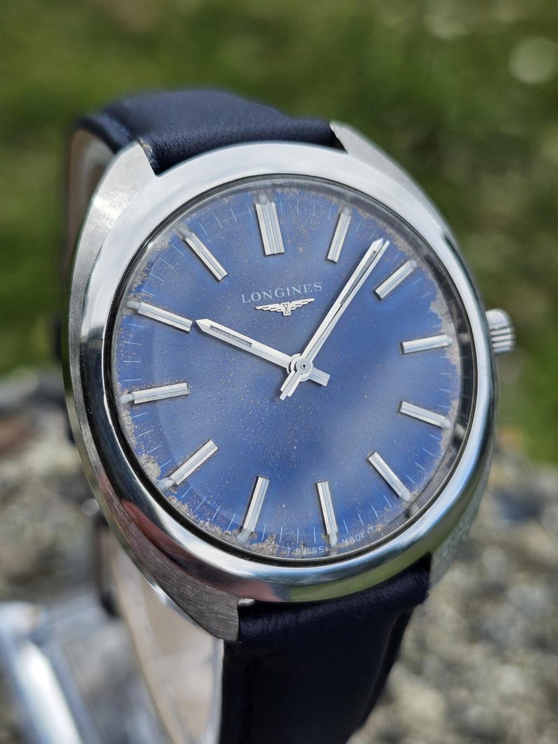 May include: A vintage Longines wristwatch with a blue dial and silver hands and markers. The watch has a stainless steel case and a dark blue leather strap. The dial features the Longines logo and the words "SWISS MADE".
