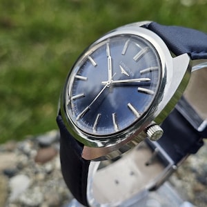 May include: A silver-toned Longines wristwatch with a dark blue face and a black leather band. The watch has silver hands and markers, and a small second hand. The watch is displayed on a clear stand.