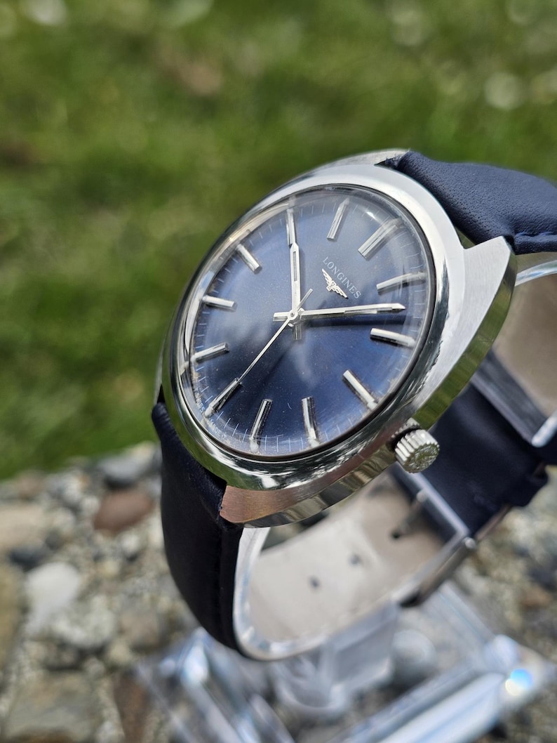 May include: A silver-toned Longines wristwatch with a dark blue face and a black leather band. The watch has silver hands and markers, and a small second hand. The watch is displayed on a clear stand.