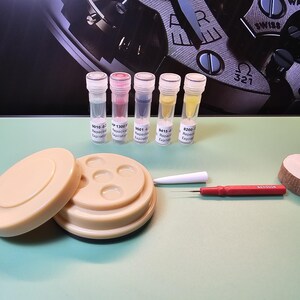 May include: An assortment of watch repair tools and components, including small vials with colored liquids, a beige parts holder, a red screwdriver, a white tool, and a wooden disc. The items are arranged on a light green surface.