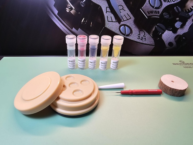 May include: An assortment of watch repair tools and components, including small vials with colored liquids, a beige parts holder, a red screwdriver, a white tool, and a wooden disc. The items are arranged on a light green surface.