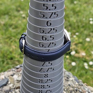 May include: A gray ring sizer with measurements from 5 to 9.5. A navy blue leather watch band is placed on the sizer. The background is blurred green grass and small rocks.