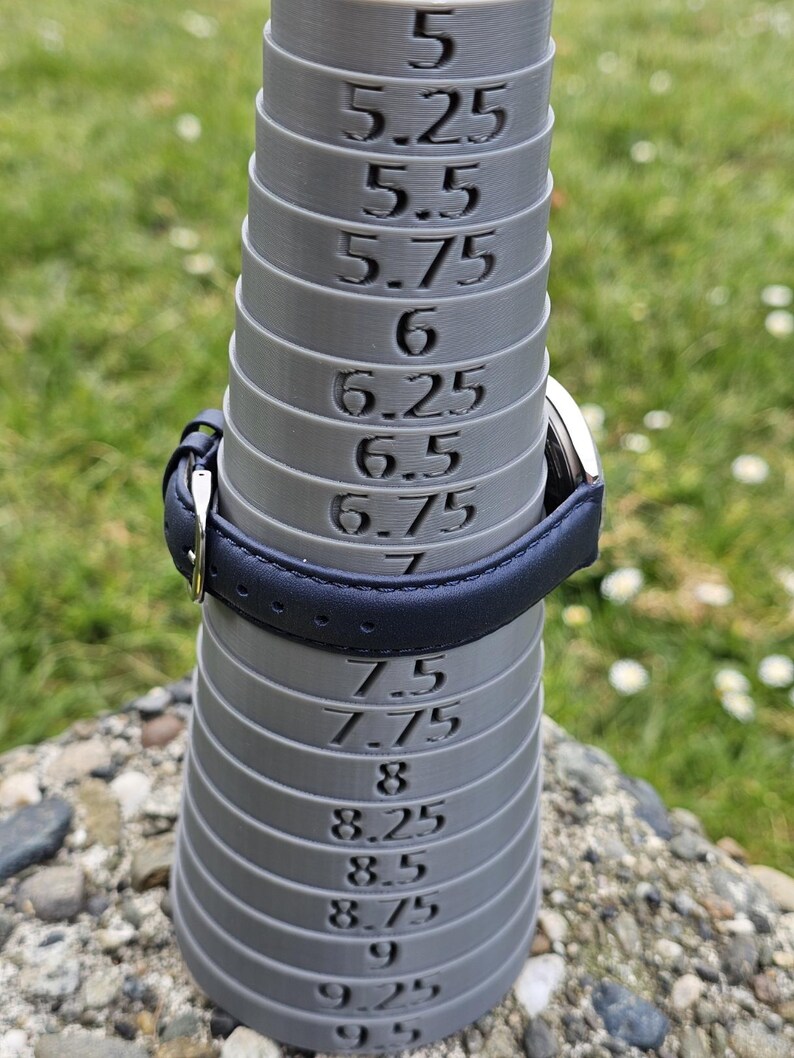 May include: A gray ring sizer with measurements from 5 to 9.5. A navy blue leather watch band is placed on the sizer. The background is blurred green grass and small rocks.