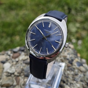 May include: A vintage Longines wristwatch with a dark blue face and silver hands. The watch has a silver case and a dark blue leather band. The watch is displayed on a clear stand against a blurred green background.
