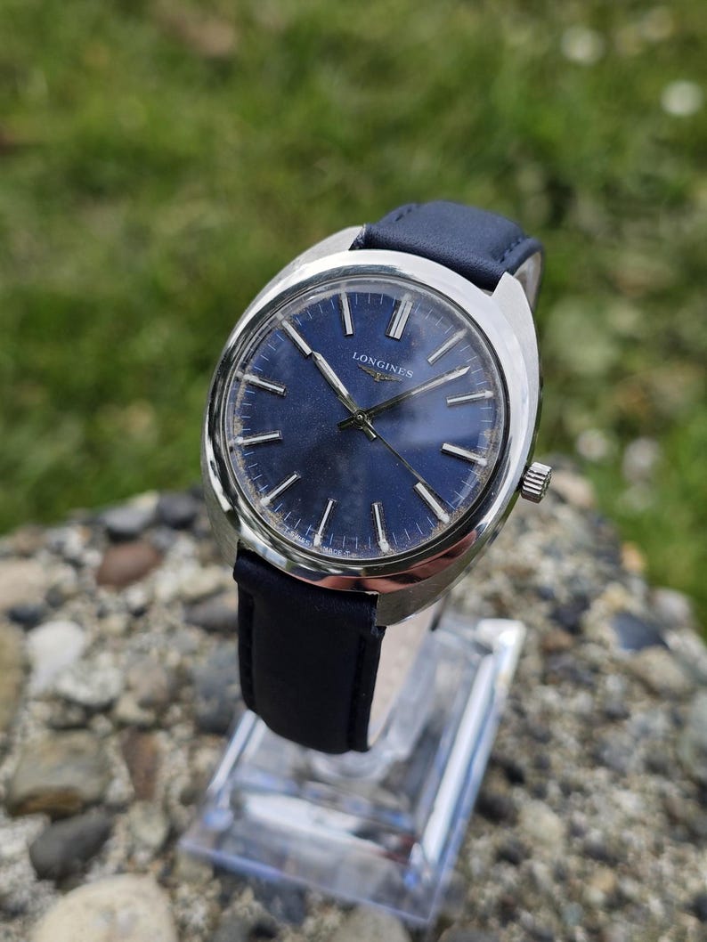 May include: A vintage Longines wristwatch with a dark blue face and silver hands. The watch has a silver case and a dark blue leather band. The watch is displayed on a clear stand against a blurred green background.