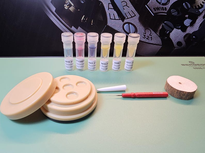 May include: A collection of watch repair tools and components. Includes small vials with colored liquids, a beige parts holder, a white tool, a red screwdriver, and a wooden disc. The background features a watch mechanism.