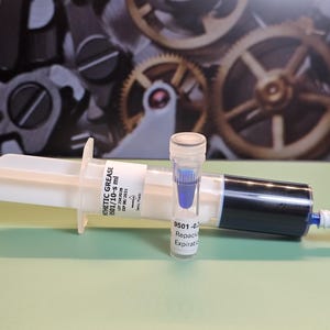 May include: A white syringe filled with black liquid and a small vial containing blue liquid. The syringe has a label that reads "SYNTHETIC GREASE". The background features gears.