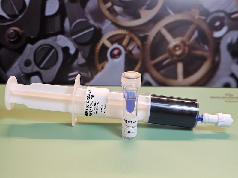 May include: A white syringe filled with black liquid and a small vial containing blue liquid. The syringe has a label that reads "SYNTHETIC GREASE". The background features gears.