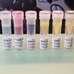 May include: Six clear plastic vials with colored liquids: blue, pink, blue, yellow, yellow, and light yellow. Each vial has a white label with text. The vials are capped with clear lids. The background is a dark, blurred surface.