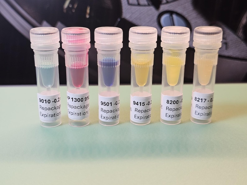 May include: Six clear plastic vials with colored liquids: blue, pink, blue, yellow, yellow, and light yellow. Each vial has a white label with text. The vials are capped with clear lids. The background is a dark, blurred surface.