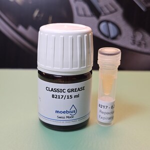 May include: A small glass bottle of "CLASSIC GREASE 8217/15 ml" with a white cap and a smaller clear vial containing a yellow liquid. The label reads "moebius Swiss Made". The background is a blurred image of a watch.